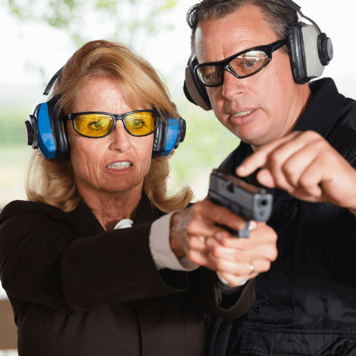 The image shows a woman wearing protective firearm accessories in the form of hearing protection and eye protection, and she's being taught by a male firearm instructor how to properly shoot her gun.