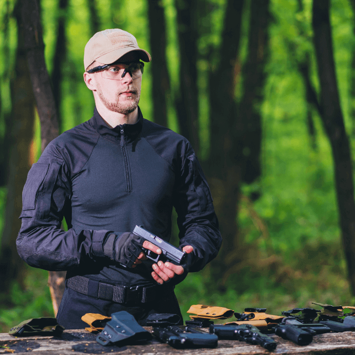 This image shows a man wearing a hat, safety goggles, and gloves, holding a gun and in front of him is a table with a variety of holsters laid out.