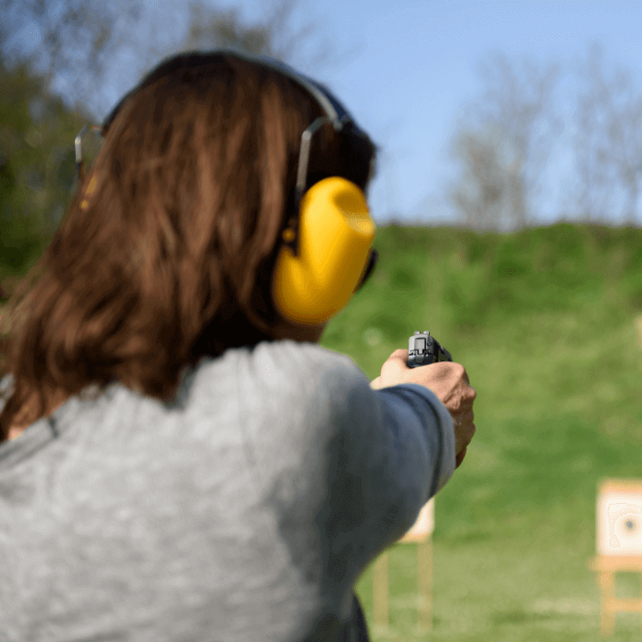 The image shows the back of a woman practicing firearm safety while holding a handgun and aiming at a target. The woman is also wearing ear protection.