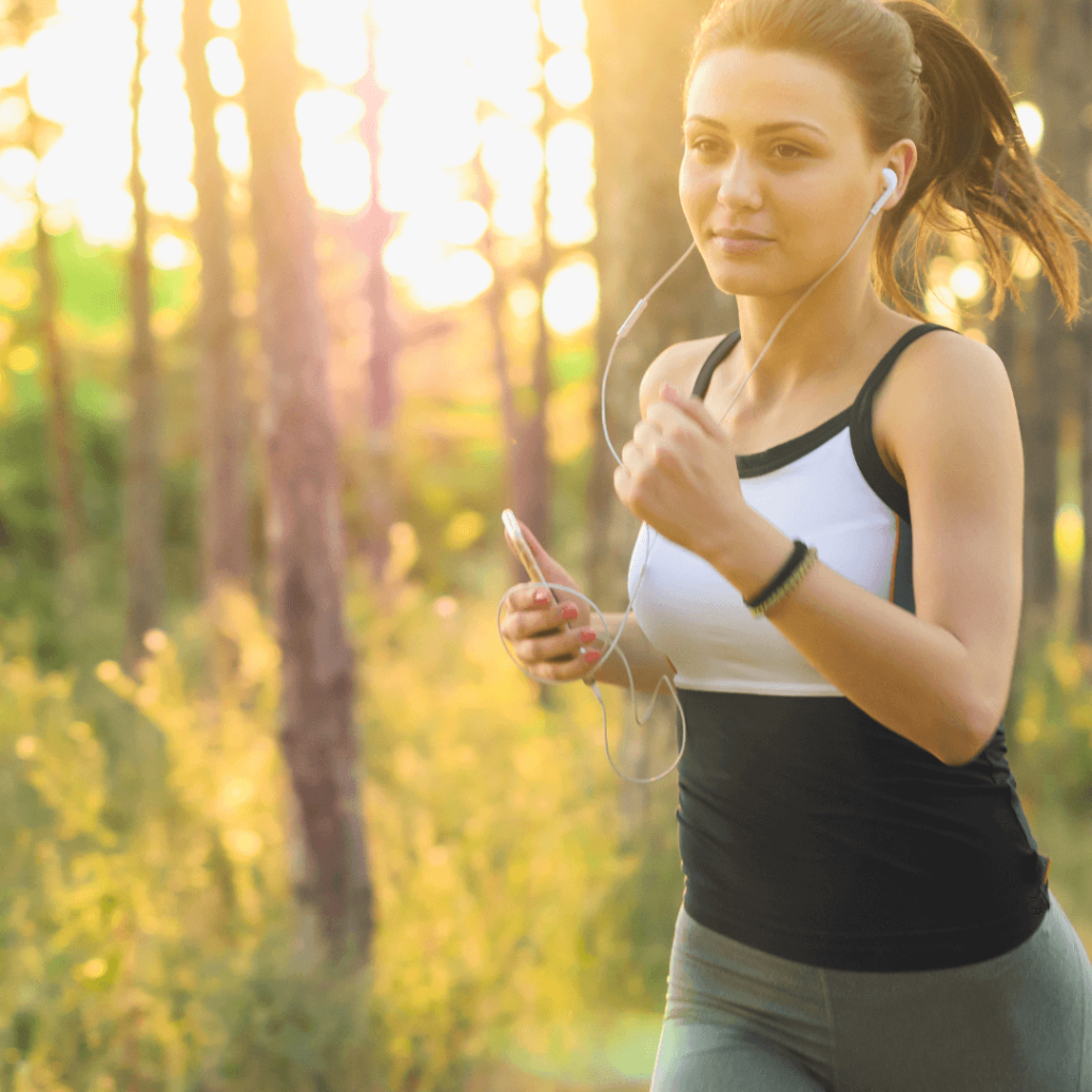 A young woman jogging, surrounded by trees, and listening to her phone with headphones in