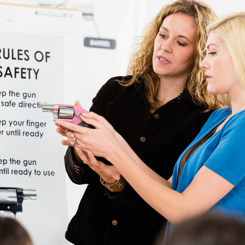 A female gun instructor teaching a younger woman how to use a gun with a set of rules listed in the background